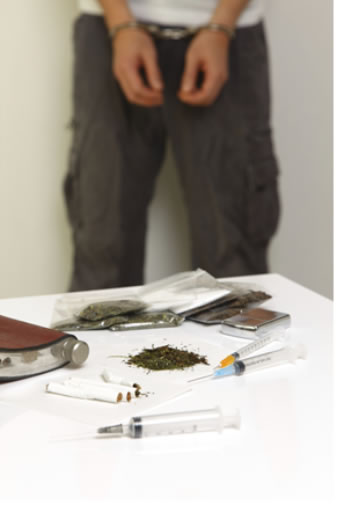 "Image is a person in handcuffs with several types of illegal drugs pictured on a table"